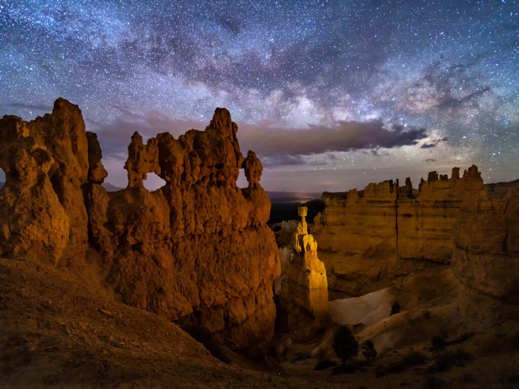 A hoodoo with small windows and Thor's Hammer against a night sky with clouds and the Milky Way